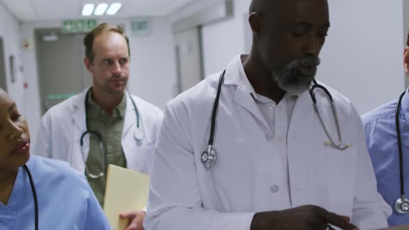 Diverse group of male and female doctors walking in hospital corridor using tablet and talking