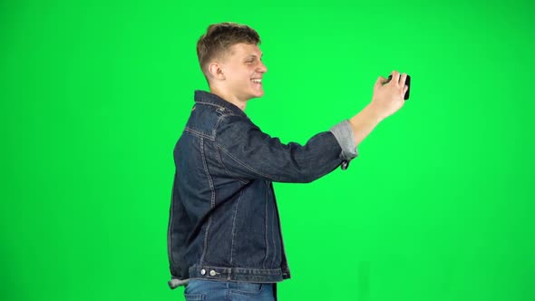 Young Man Goes and Takes a Selfie with Smartphone on Green Screen at Studio. Slow Motion. Side View