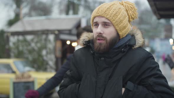 Handsome Caucasian Man in Yellow Hat Standing on Winter Street As His Beautiful Girlfriend or Wife