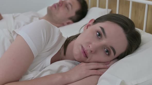 Sad Woman Looking at Camera Next to Sleeping Partner in Bed