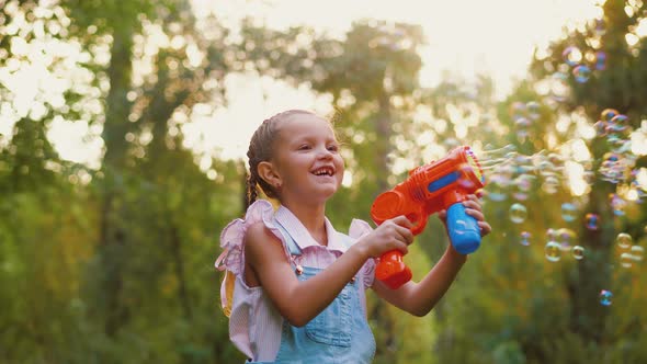 Happy Little Girl Blowing Soap Bubbles Playing in Park Outdoors with Toy Gun