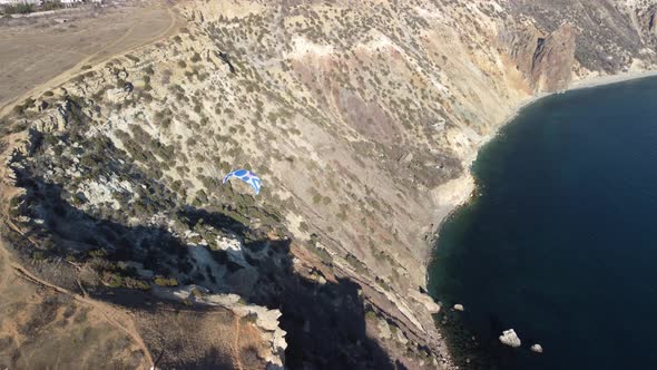 Aerial Drone View of a Man Flying a White and Blue Paraglider Over a Hill and Trees to the Sea Waves