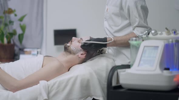 Side View of Relaxed Handsome Man Lying in Beauty Salon As Unrecognizable Beautician Using LED Light