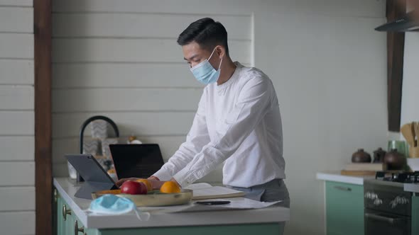 Side View of Concentrated Chinese Man in Covid19 Face Mask Typing on Tablet Messaging Online