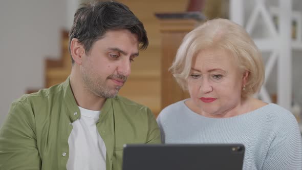 Portrait of Confused Senior Woman and Patient Adult Man Talking Pointing at Tablet