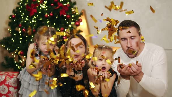 Young Happy Family of Four Sitting By the Christmas Tree and Blowing Golden Confetti