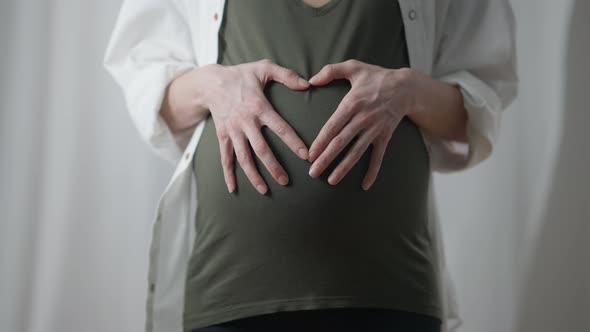 Closeup Pregnant Woman Gesturing Heart Shape on Belly Standing Indoors