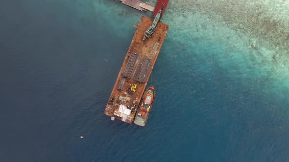 Crane loading pipes on flat industrial boat surrounded by clear transparent indian ocean with coral