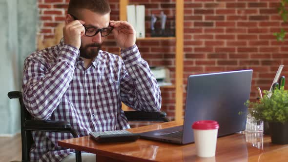 Hipster Man with Disabilities Sits on Wheelchair at Table with Laptop Uses It Counts on Calculator