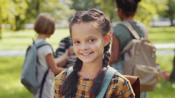 Portrait of Cheerful Schoolgirl with Backpack Posing for Camera in Park