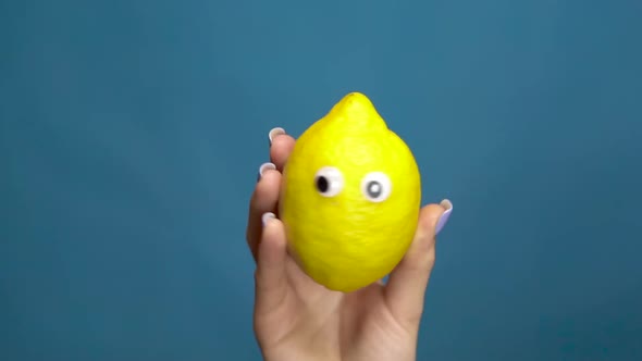 Lemon with Eyes in a Woman Hand Close-up. Lemon Shakes and Twists Eyes on a Blue Background. Slow