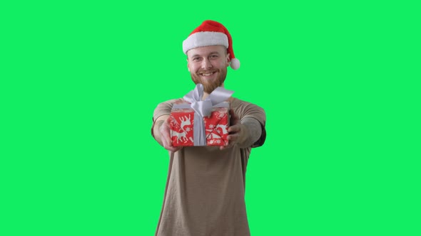 Young Handsome Bearded Tattooed Guy in Green T-shirt with Red Christmas Hat Is Giving a Gift To the
