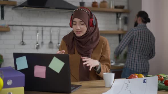 Young Confident Middle Eastern Woman in Headphones Conferencing Online Drinking Coffee in Home