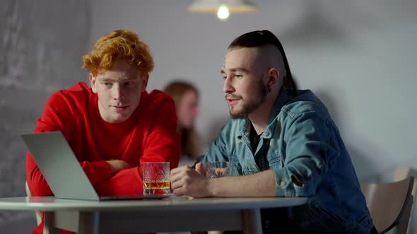 Two Handsome Young Men Sitting in Cafe with Alcohol Using Laptop and Talking in Slow Motion