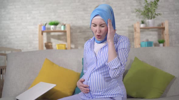 Young Pregnant Muslim Woman in a Hijab Will Experience Headaches Sitting on the Couch