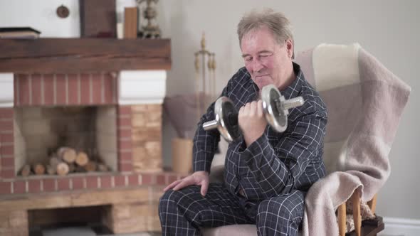 Elderly Caucasian Man Lifting Dumbbell As Sitting in Armchair at Home, Active Old Retiree Exercising