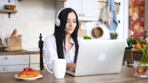 Smiling Young Woman Using Laptop In The Kitchen At Home Wearing Headphones