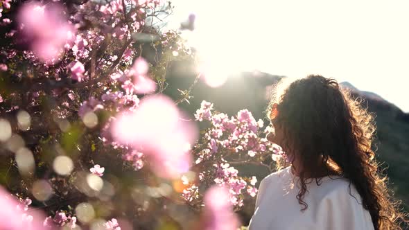 A Young Woman with Long Curly Hair Enjoys a Blooming Spring Garden at Sunset in the Mountains