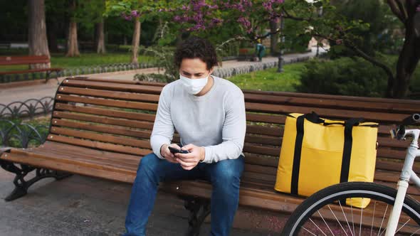 Portrait of Caucasian Delivery Man Sitting on Bench in Central Park and Using Smartphone with Yellow