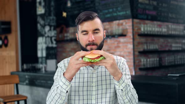 Pleasant Hipster Stylish Young Man Biting Juicy Appetizing Burger Enjoying Taste Looking at Camera