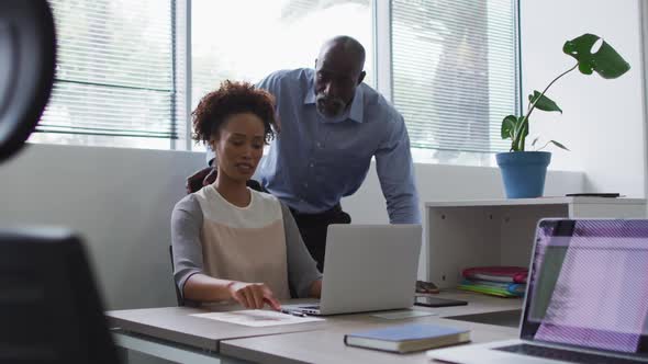 Diverse businessman and businesswoman talking, woman using laptop in office