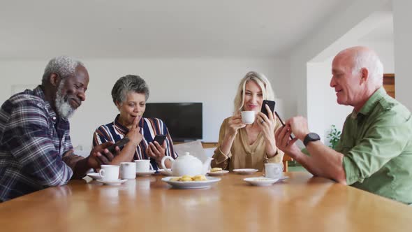 Two diverse senior couples sitting by a table drinking tea using smartphones at home