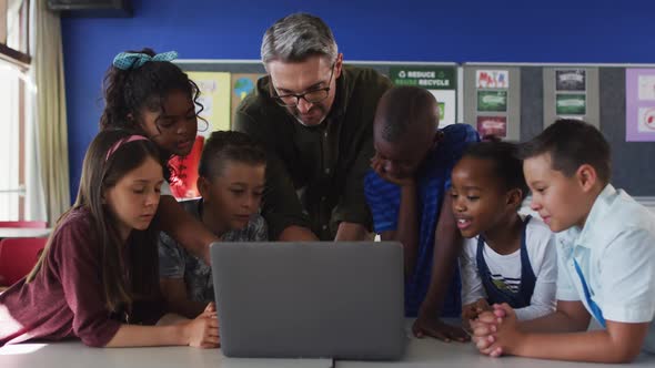 Diverse male teacher and group of schoolchildren looking at laptop