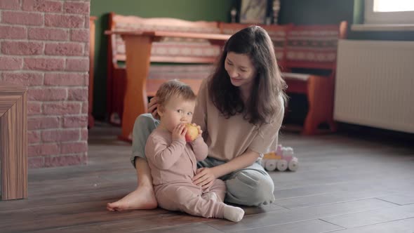 Happy Beautiful Woman Sitting at Kitchen Table Having Breakfast and Feeding Her Baby Daughter