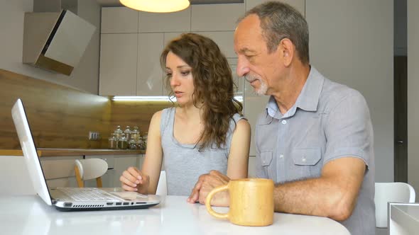 Attractive Young Daughter and Senior Father are Using Laptop Sitting in Modern Kitchen in Apartment