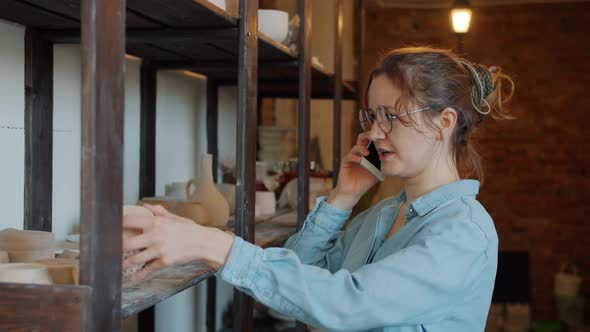 Cheerful Girl Talking on Mobile Phone Holding Hand-made Ceramics in Pottery Studio