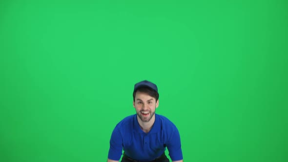 Cheerful Man Courier in Uniform Stands with a Cardboard Box in His Hands on a Green Background