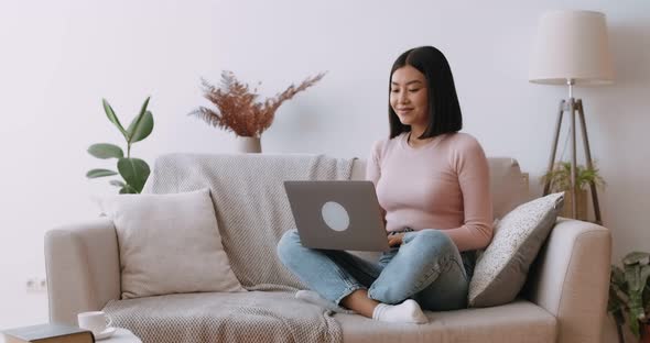 Young Asian Woman Networking on Laptop at Home, Freelancing Online at Home, Sitting on Couch
