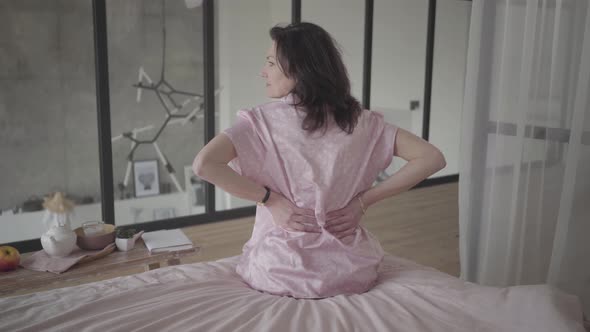 Serious Adult Caucasian Woman Rubbing Back As Sitting on Bed Indoors. Back View of Gorgeous Brunette