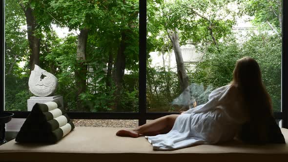 Young Woman Lying on Couch Near Window with Greenery in the Background