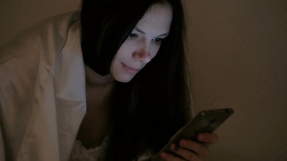 Young Beautiful Woman Brunette Reads a Book in Her Mobile Phone in Bed Before Sleeping and Smiles
