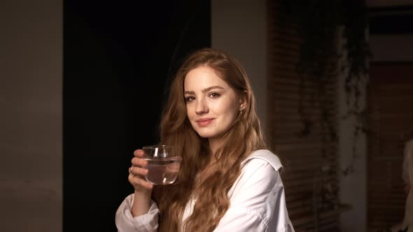 Red-Haired Girl In White Shirt Holds Glass Of Water In Her Apartment.