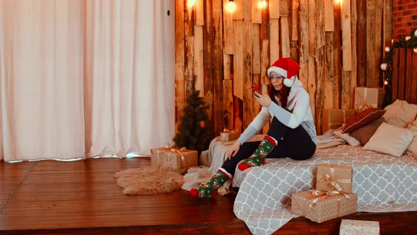 Young Woman in Santa Claus Hat Browses Smartphone Sitting on Bed