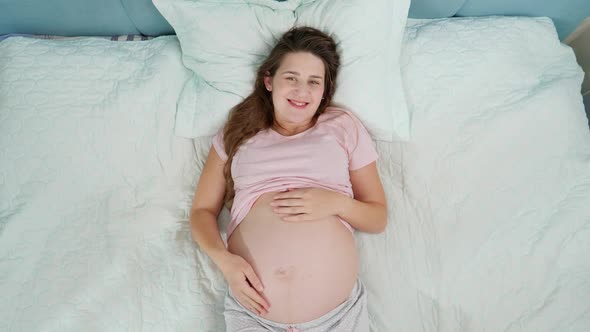 Top View of Happy Smiling Pregnant Woman Enjoying Pregnancy Lying on Bed and Stroking Her Big Belly