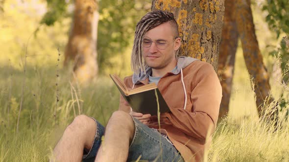 A man in casual clothes and glasses reads literature under a tree.