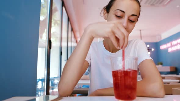 Pretty Young Tanned Woman in White T-shirt Is Sitting in Cafe and Drinking a Cold Lemonade or