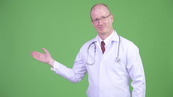 Happy Mature Bald Man Doctor Showing Something Against Green Background