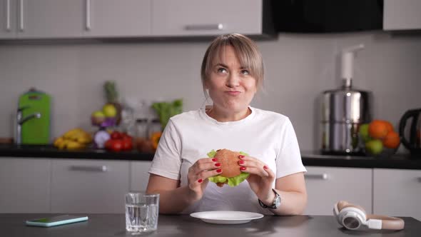 Hungry Woman Eating Burger Sitting in Kitchen