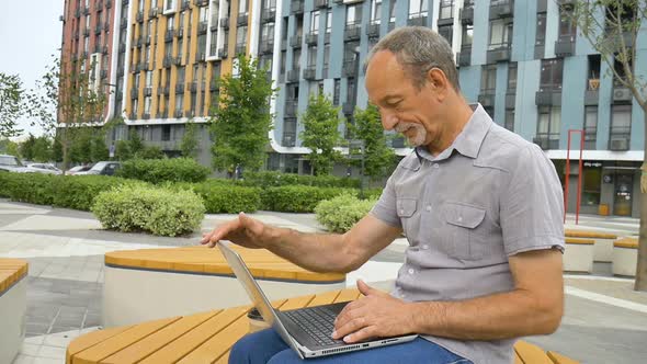 Trendy Mature Man is Working Outside with Laptop Sitting at the Bench Near New Modern Residential