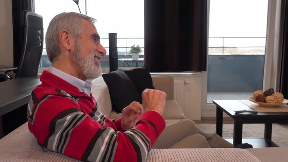 A Happy Elderly Man Sits on a Couch in an Apartment and Dances with a Smile - Side Closeup