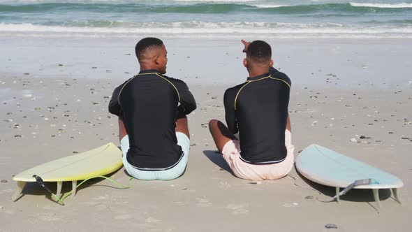 African american teenage twin brothers sitting by surfboards on a beach talking