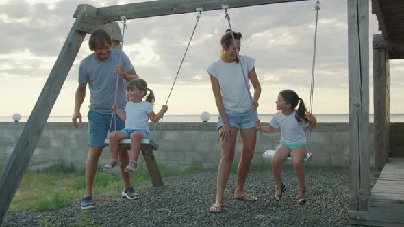 Happy Family Rolls Young Children on a Swing Outdoors at Sunset