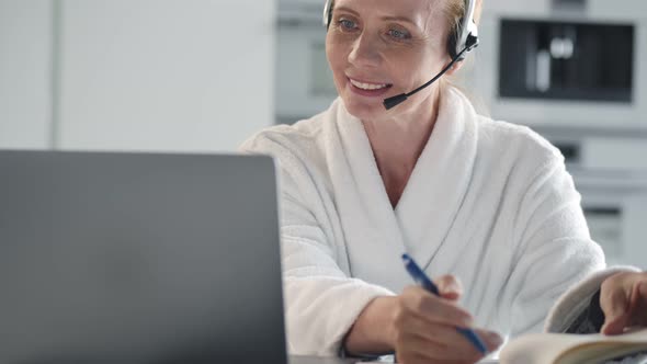 Mature Woman with Headset and Laptop Having Video Conference and Taking Notes at Home Office