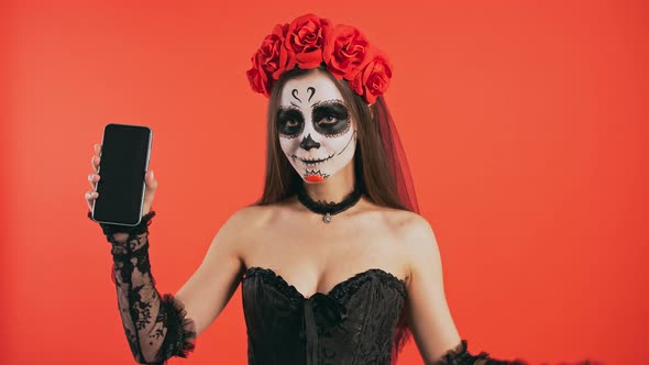 Day of Dead Woman with Skull Makeup Holding Smartphone Pointing at Its Screen By Forefinger and