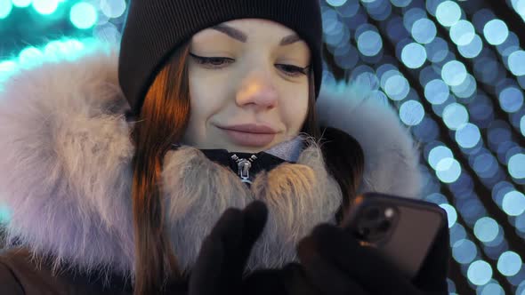 Young Beautiful Woman Uses the Phone Against the Background of the Central Christmas Tree Lights and