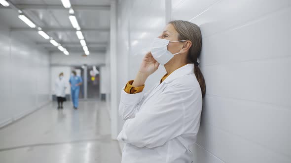 Sad and Depressed Aged Female Doctor Having Migraine in White Uniform Standing in Corridor